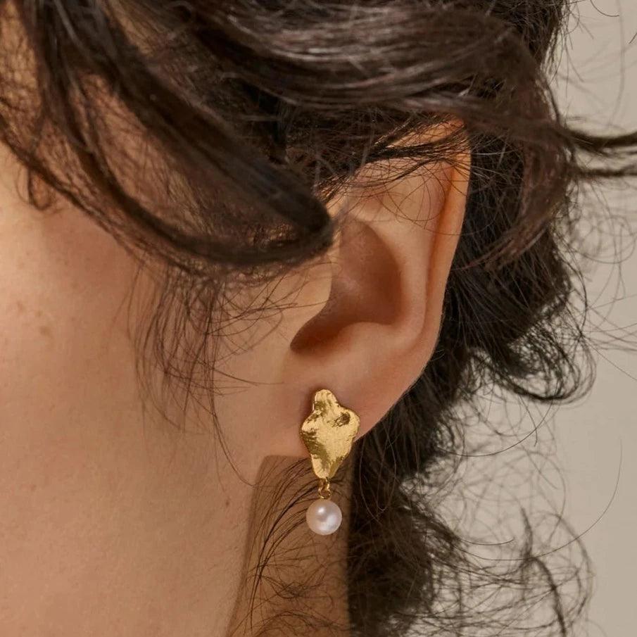 enamel-caia-small-örhängen-e374gm visas på örat, matt guldlook med hängande vit pärla. Modell E374GM