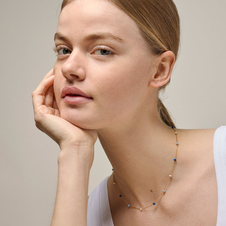 enamel-lola-bold-halsband-marin-i-förgylld-sol n137g-marin visas på modell, finguldkedja med blå och ljusa pärlor, modell n137g-marin