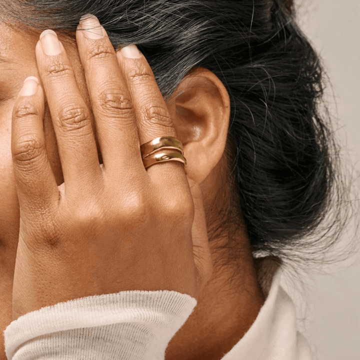 Enamel Sandra Statement Ring guldpläterad visas på kvinnans hand nära örat, glänsande gyllene yta. Modell R92G-R92G-001.