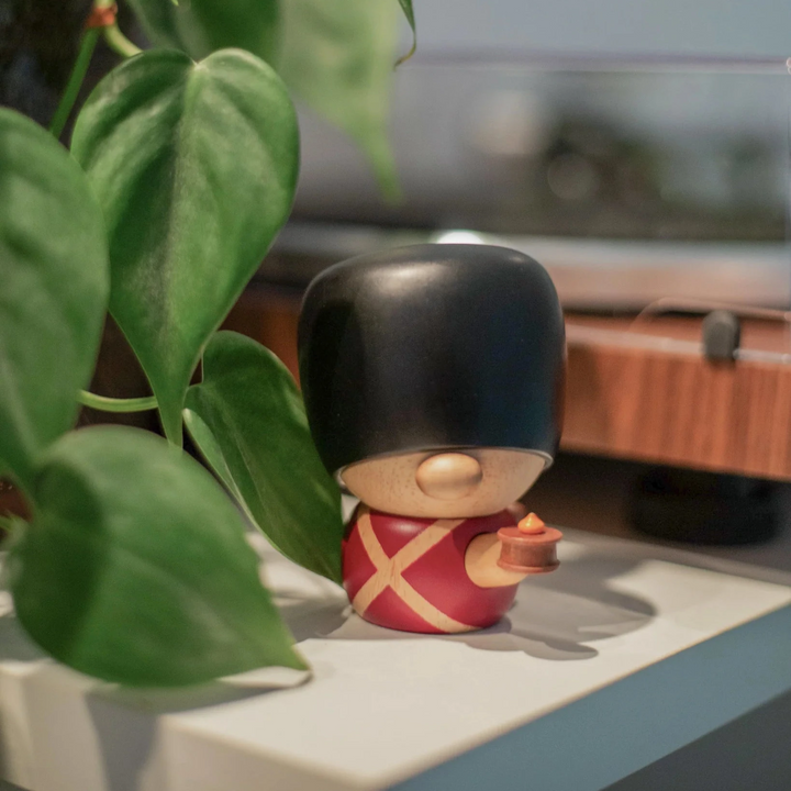 Fablewood HC Andersens Fyrtøjet-8005-1 träfigur av vakter i röd uniform och svart björnskinnshatt på hylla vid grön växt. Modell 8005-1