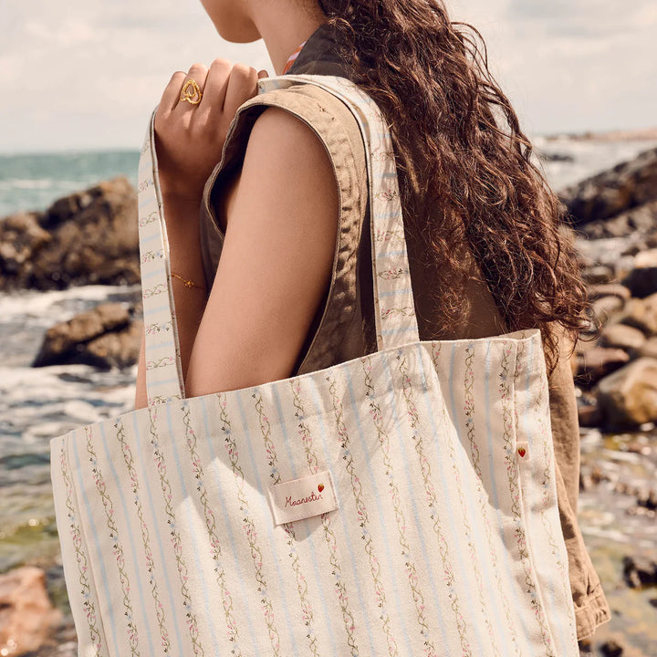 maanesten-canvas-totebag-sommar-ränder-blå bärs utomhus vid kusten, ljusblå ränder och broderade detaljer. Modell 4121-one size