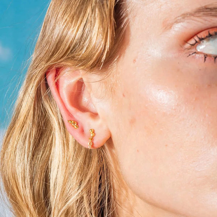 Vincent Pendelörhängen Guldpläterade visas på örat, små kulor i guld ser ut under klar himmel. Modell VJ177-UEG-2